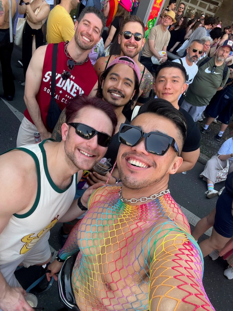 A group of seven men smiling and posing for a selfie at a vibrant outdoor event, wearing colorful attire, including a rainbow net shirt and sunglasses, with a crowd in the background.