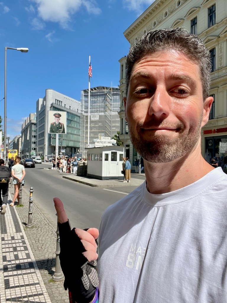 A selfie of a man in a white shirt standing on a busy street, pointing towards a military-themed poster on a nearby building, with a view of traffic, pedestrians, and a clear blue sky.