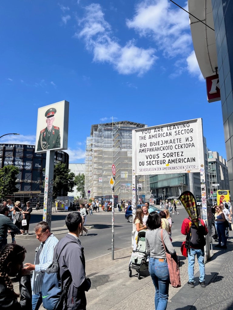 A busy street scene featuring a sign that reads 'YOU ARE LEAVING THE AMERICAN SECTOR' in multiple languages, alongside a large poster of a soldier. There are pedestrians and cars in the foreground, with buildings and a blue sky in the background.