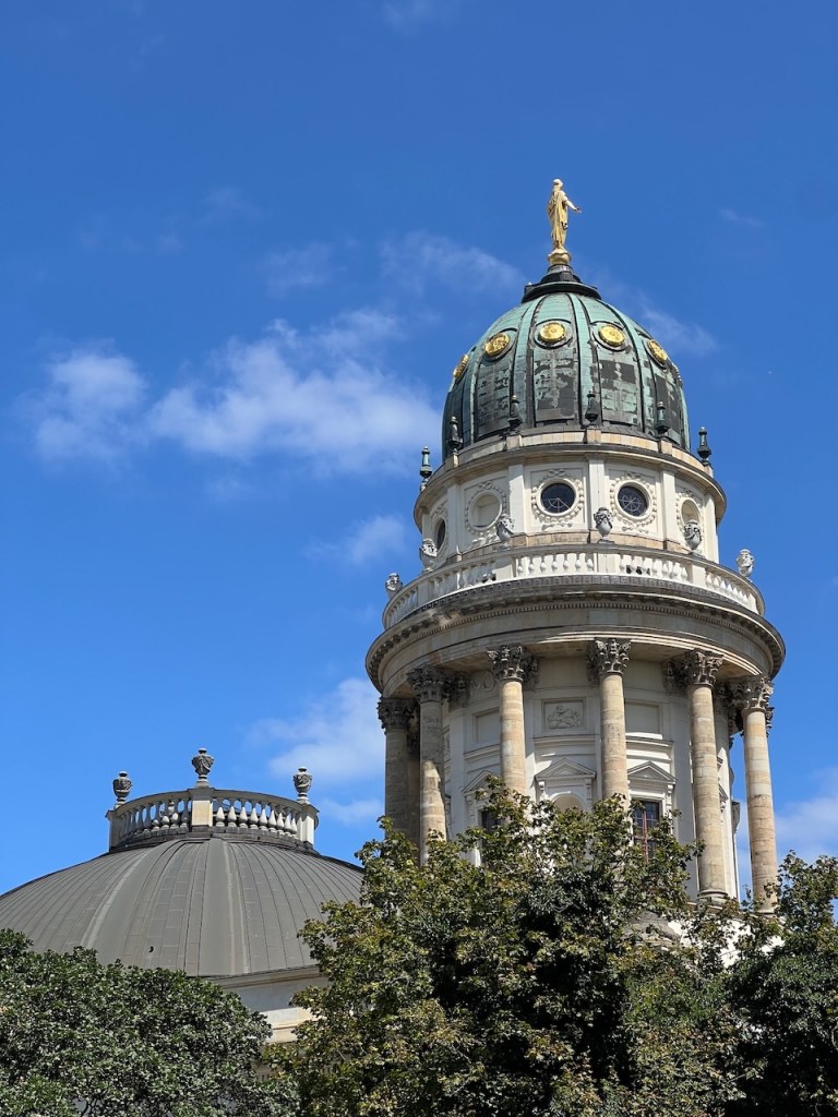 Close-up of a historical dome with a statue atop, surrounded by trees and a bright blue sky.