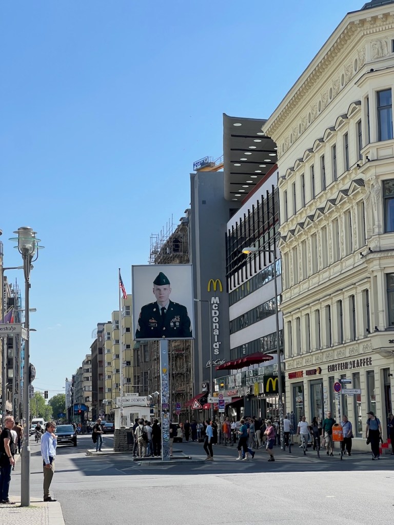 A busy city street with people walking, featuring a large billboard displaying a military officer's portrait. The McDonald's logo is visible on a nearby building, along with various shops and structures.