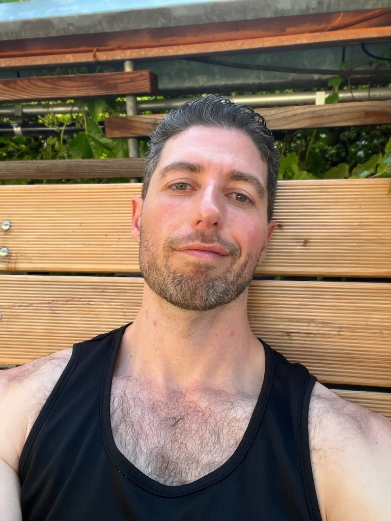 A man with short dark hair and a trimmed beard is sitting on a wooden bench, wearing a black tank top, and looking directly at the camera with a relaxed expression.