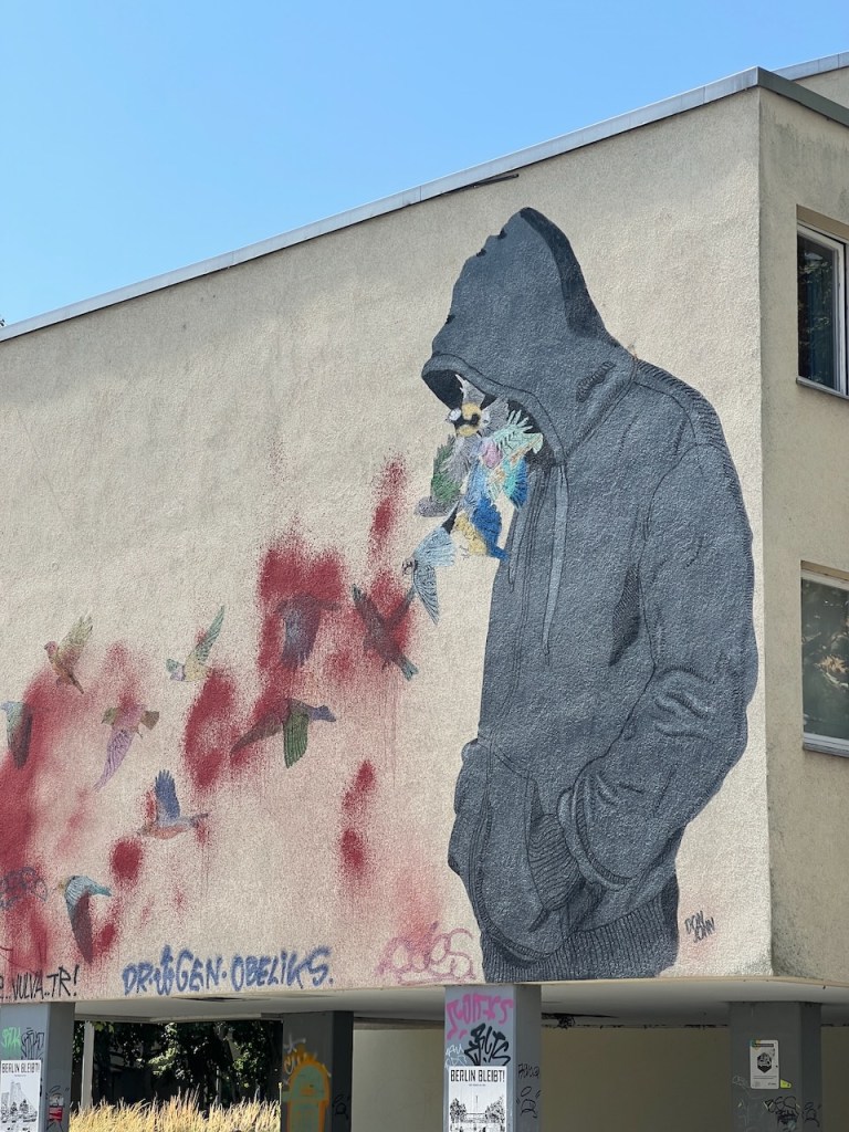 A large mural depicting a figure wearing a hoodie with floral elements emerging from the hood, set against a background of red splashes and birds in flight.