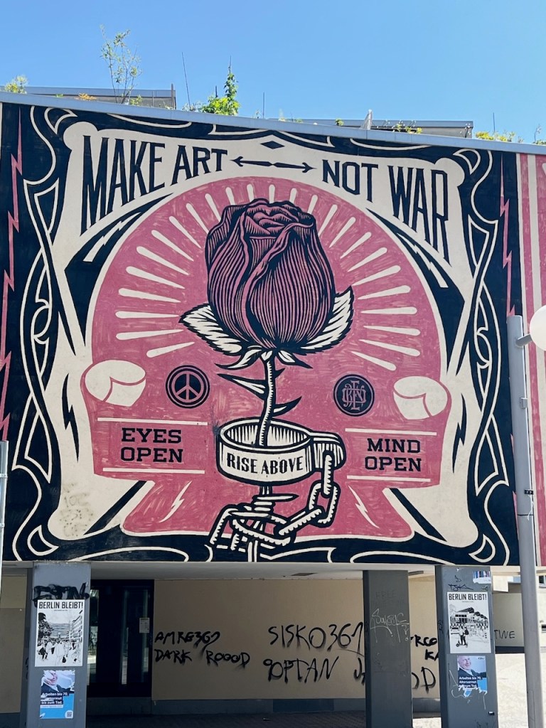 Mural featuring a large rose with text that reads 'MAKE ART NOT WAR,' 'EYES OPEN,' 'MIND OPEN,' and 'RISE ABOVE.' The design includes peace symbols and striking graphic elements.