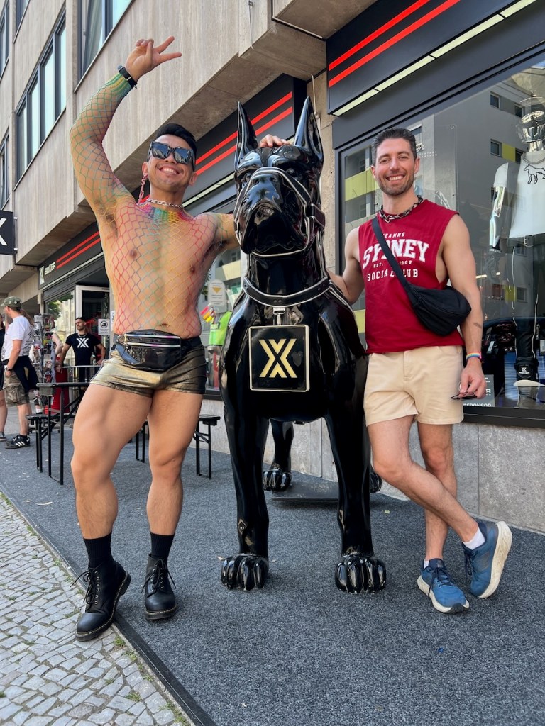 Two men posing in front of a large black dog statue on the sidewalk. One man is wearing colorful mesh clothing and shorts, while the other is dressed in a red tank top and shorts.