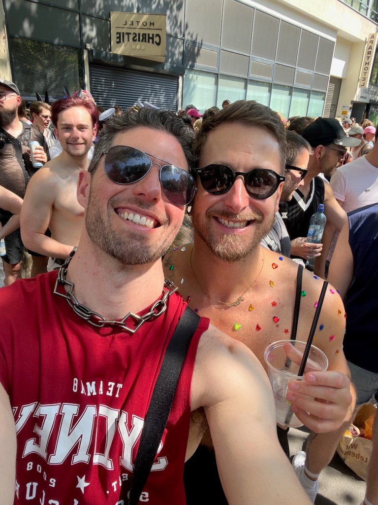 Two smiling men taking a selfie at an outdoor event, one wearing a red sleeveless shirt and a chain necklace, and the other adorned with colorful decorations on his chest, with a crowd and buildings in the background.