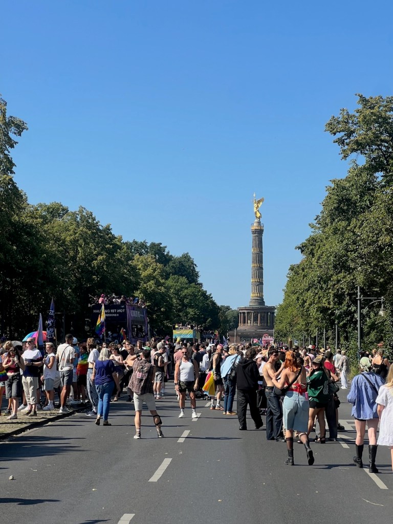 A vibrant street scene filled with a diverse crowd celebrating at a parade, with the Victory Column in the background under clear blue skies.