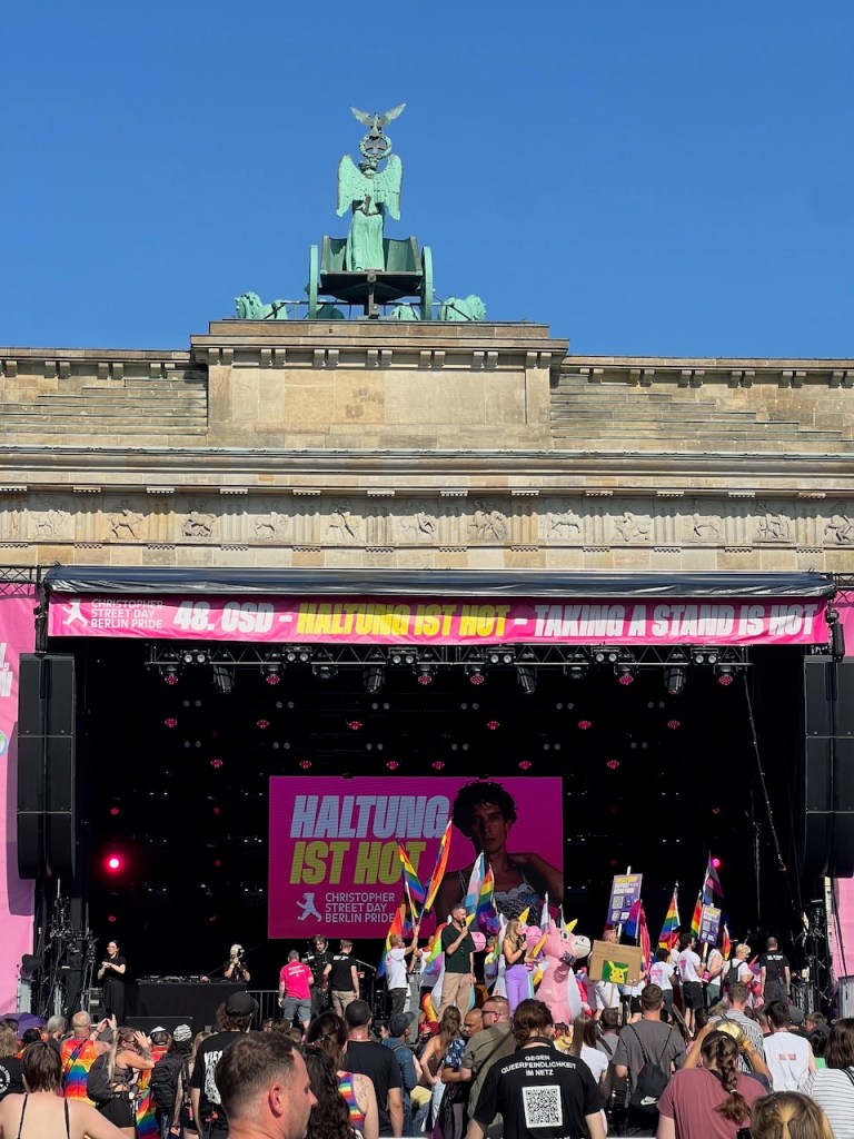 A vibrant scene from Berlin Pride with a large stage displaying 'HALTUNG IST HOT' in bold letters, surrounded by a crowd holding rainbow flags and celebrating. The iconic Brandenburg Gate is visible in the background under a clear blue sky.