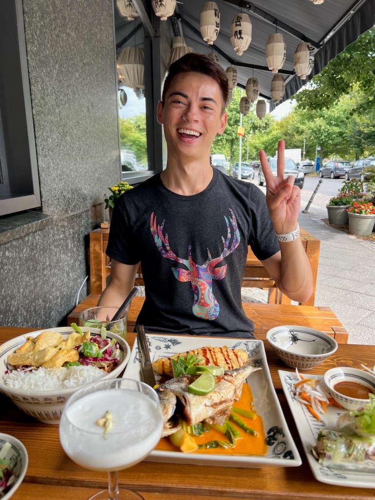A young man sitting at a restaurant table, smiling and giving a peace sign. He is wearing a black t-shirt with a colorful deer design. The table is filled with several dishes, including grilled fish, rice, salad, and a cocktail.
