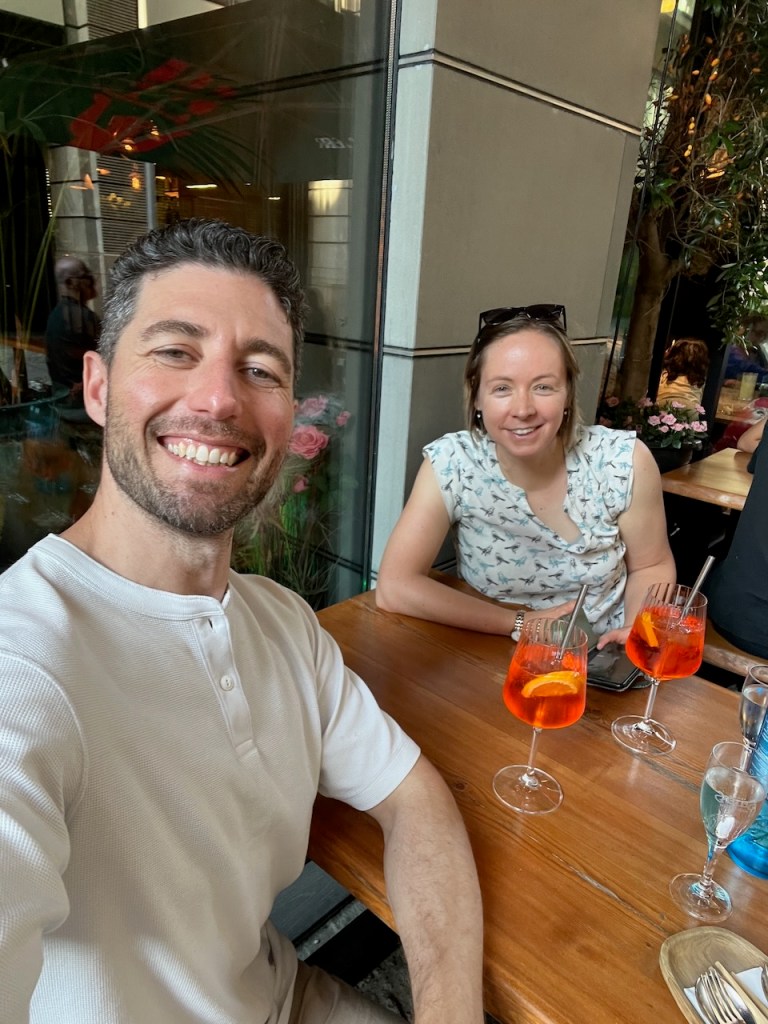 A man and a woman smiling at the camera while sitting at a restaurant table with drinks in front of them.