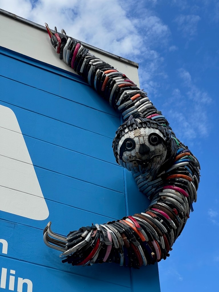 A large, imaginative sculpture of a sloth made from various colorful materials, including metal and plastic, mounted on a blue wall, reaching out with one arm.