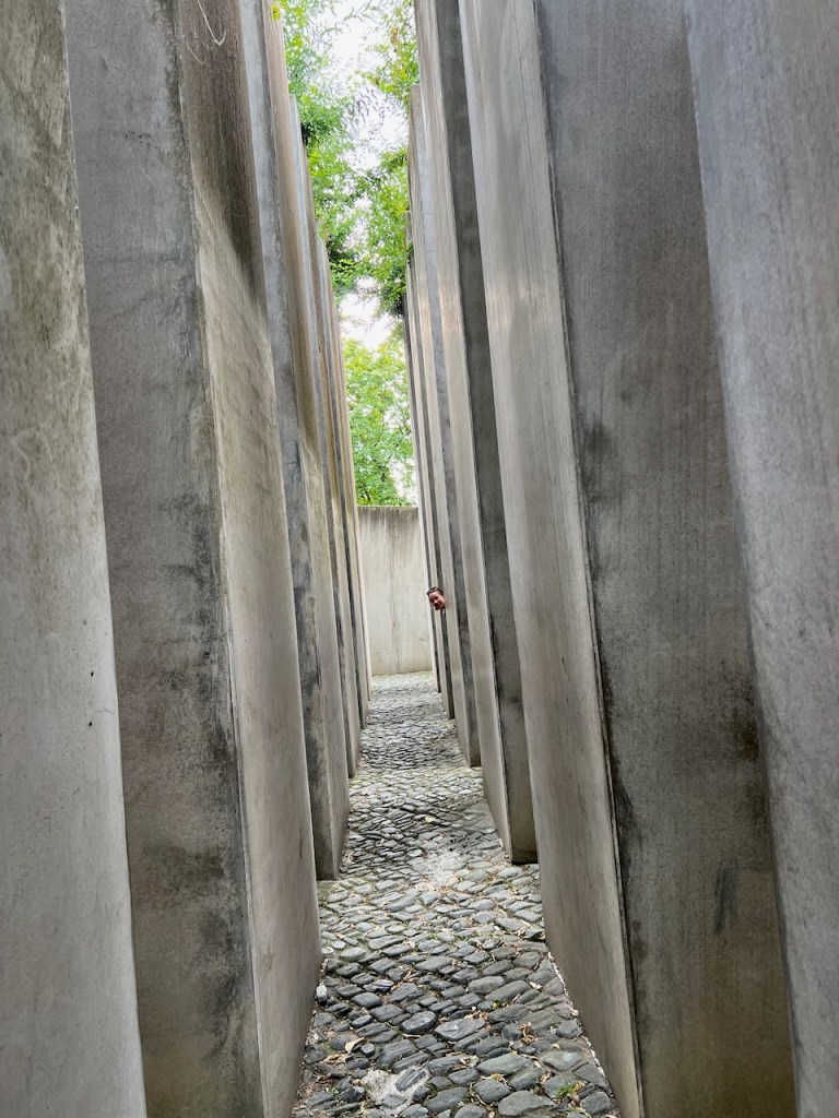 Narrow pathway between tall concrete slabs in a monument, featuring a cobblestone surface and greenery visible above.