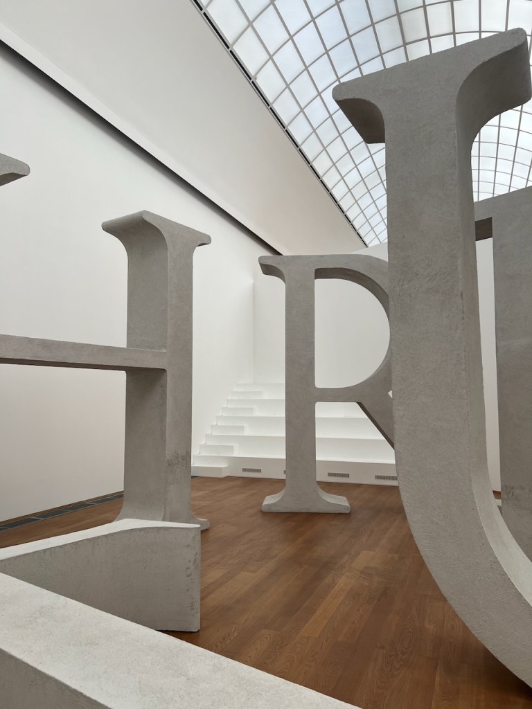 Large sculptural letters 'H', 'R', and 'U' in a modern art gallery with a glass ceiling and wooden flooring.